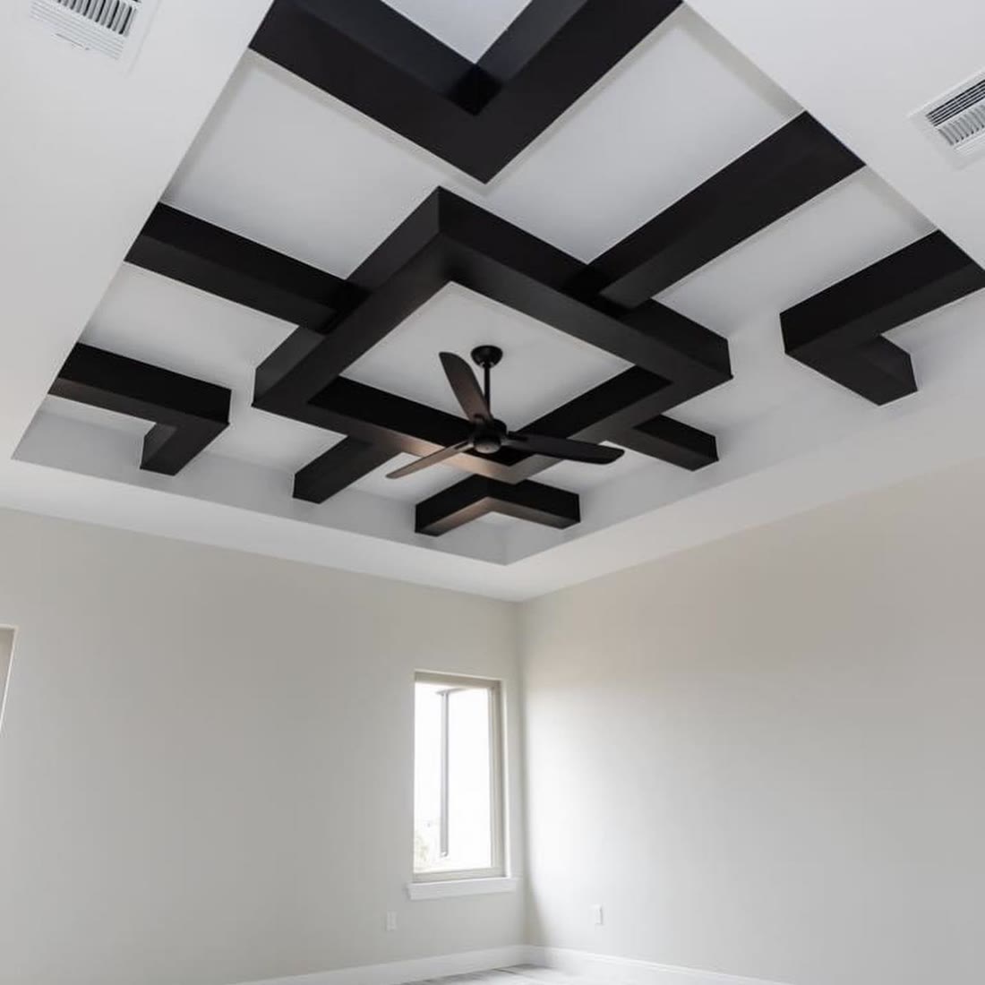 A white room with a geometric ceiling of black beams around a black ceiling fan.