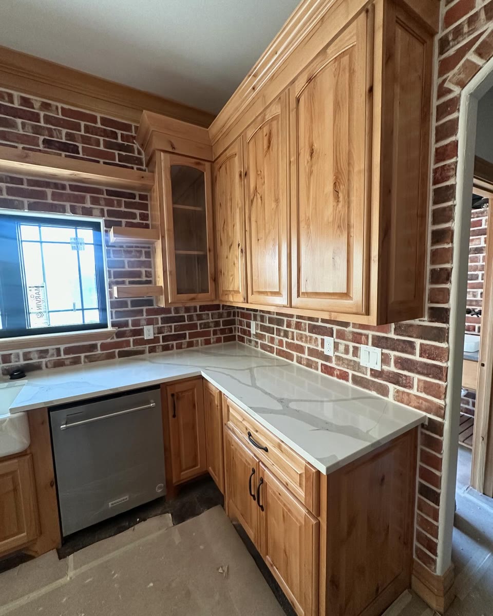 Knotty wood kitchen cabinets against a red brick wall, a white stone counter below.