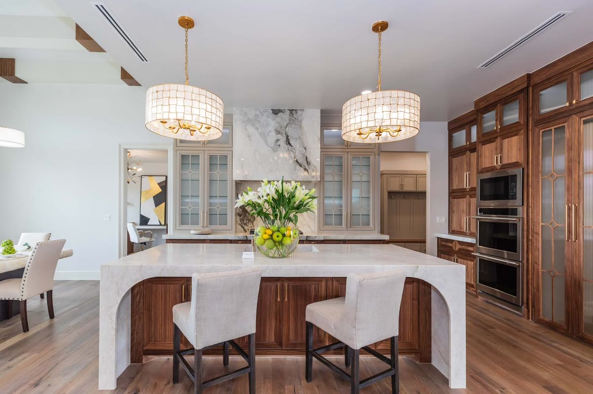 A white waterfall island with two white stools, stained cabinets and wall ovens behind it.