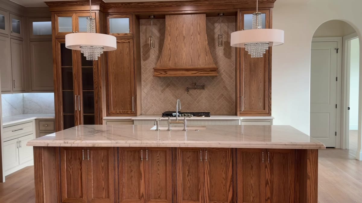 A kitchen with stained lower cabinets, a carved wood range hood and two chandeliers.