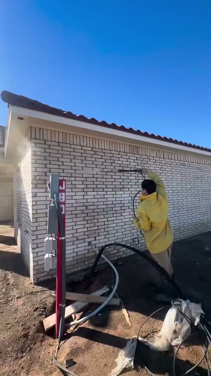 A painter in a yellow rain jacket spraying a brick wall white, the hose and pump beside the painter.
