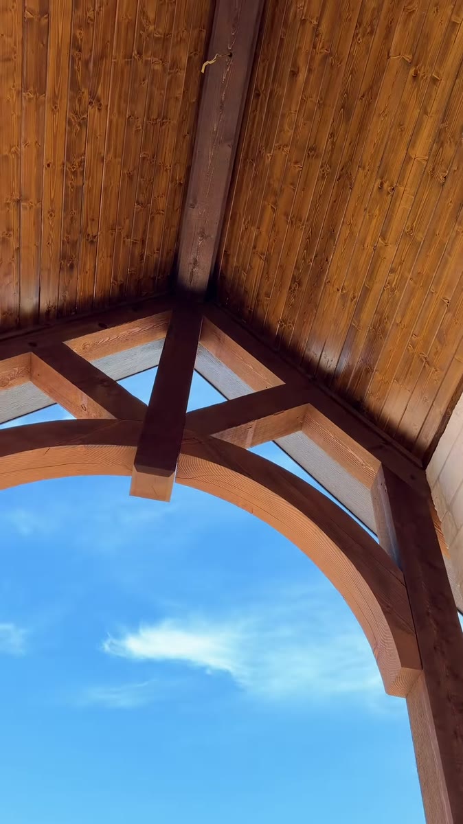 Looking up through a stained timber arch and trusses at a blue sky.